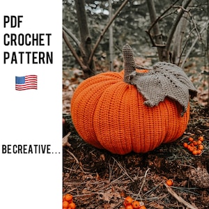 May include: A crocheted orange pumpkin with a brown crocheted stem and leaf. The pumpkin is sitting on a bed of brown leaves and twigs.