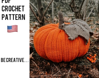 May include: A crocheted orange pumpkin with a brown crocheted stem and leaf. The pumpkin is sitting on a bed of brown leaves and twigs.