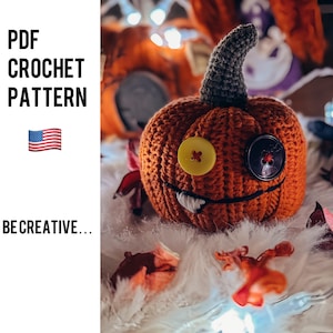 Puede incluir: Una calabaza naranja tejida con un tallo gris, ojos de botón amarillos y negros y una boca cosida con dientes blancos. Se muestra el texto "PDF CROCHET PATTERN" y "BE CREATIVE...". También se muestra una bandera estadounidense.