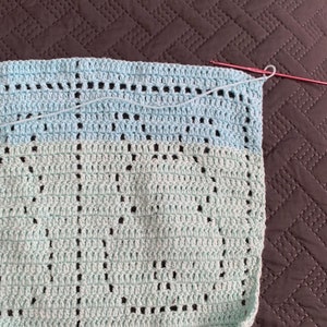 May include: A light blue crochet blanket with a pattern of circles. The blanket is partially finished and has a pink crochet hook on the right side.