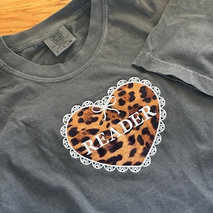 May include: Dark grey t-shirt featuring a leopard print heart design with white lace trim and the word "READER". The shirt is made of a soft material. The tag says "COMFORT COLORS".