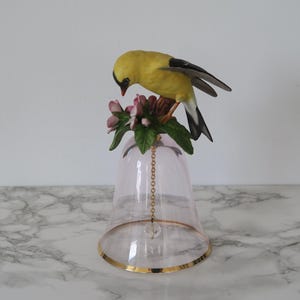 May include: A decorative bell featuring a translucent pale pink glass body with a gold-toned rim. The handle is topped with a hand-painted porcelain yellow bird perched on a cluster of pink flowers and green leaves, with a gold chain clapper.