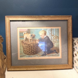 May include: A framed vintage-style print featuring a young child with blonde hair kneeling beside a woven wicker basket. A small tan pug stands on top of the basket. The artwork is enclosed in a textured gold-toned frame with a gray mat border.