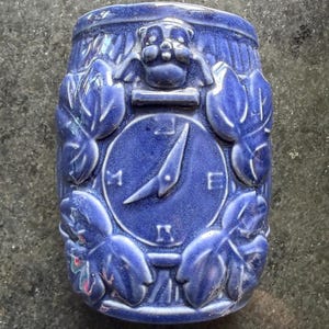 May include: A cylindrical blue ceramic vessel featuring a relief design of a compass face with cardinal directions marked as N, S, E, and W. The surface is decorated with sculpted leaf motifs surrounding the compass and a small animal figure at the top.