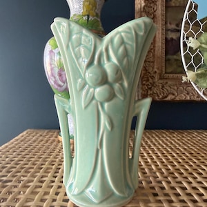 May include: A light green ceramic vase with a stylized fruit and leaf design. The vase has a flared top and handles on either side. The vase is sitting on a woven wicker surface.