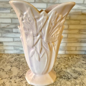 May include: A light pink ceramic vase with a flared top and a pedestal base. The vase features embossed floral and leaf designs. The vase is a soft, muted pink color and is likely vintage.