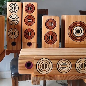 Puede incluir: Un conjunto de altavoces de madera hechos a mano de varios tamaños. Los altavoces presentan diseños circulares y concéntricos con detalles en madera clara y oscura. Los altavoces están dispuestos sobre una superficie de vidrio.