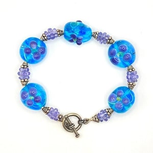 Puede incluir: Una pulsera artesanal con vibrantes cuentas de vidrio azul con diseños florales morados. La pulsera incluye cuentas moradas más pequeñas y detalles metálicos plateados, asegurada con un cierre de palanca. Una hermosa pieza de joyería.