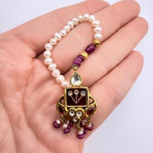 May include: A handcrafted pendant necklace featuring a square gold-toned pendant with a red center and clear and red gemstones. The pendant is connected to a strand of white pearls and red and gold beads.