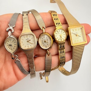 May include: A collection of five analog wristwatches with metal bands. The watches feature various case shapes including oval, square, and round, with silver and gold-tone finishes. Each watch has a dial with hour markers and hands for timekeeping.