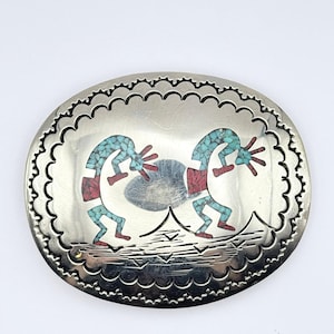 May include: Oval silver belt buckle with a scalloped edge. The buckle features two turquoise and red Kokopelli figures. The figures are dancing. The buckle is a Southwestern style.