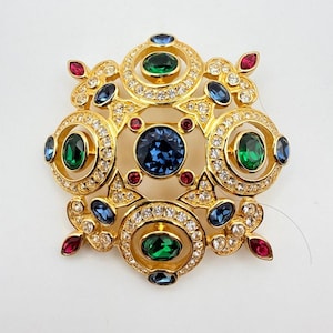 Könnte beinhalten: Eine goldfarbene Brosche mit einem zentralen, dunkelblauen, runden Kristall, umgeben von vier smaragdgrünen, ovalen Kristallen in runden Fassungen. Das Schmuckstück ist mit kleineren blauen und roten Kristallen sowie klaren Strasssteinen verziert.