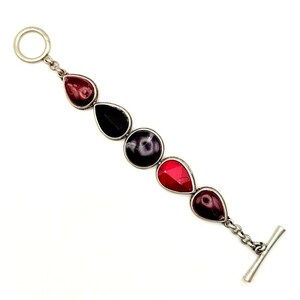May include: A silver-toned toggle bracelet featuring five distinct gemstones in a linear arrangement. The stones include teardrop-shaped red and dark maroon gems, a solid black teardrop, and a central round dark purple stone, all set in metal.