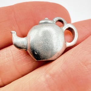 May include: A small, silver-toned metal charm shaped like a teapot. The charm features a rounded body, a curved spout on the left side, a small lid detail on top, and a circular handle on the right side. The surface has a matte, metallic finish.