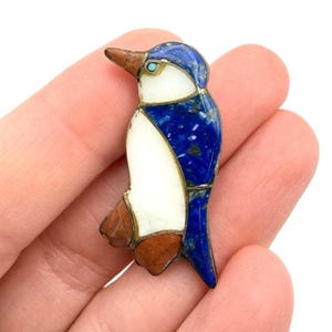 Puede incluir: Broche con forma de pájaro con un diseño azul, blanco y marrón. El cuerpo del pájaro es principalmente blanco, con detalles azules en la cabeza y las alas. El pico y las patas son marrones. Una pequeña piedra turquesa está engastada cerca del ojo.