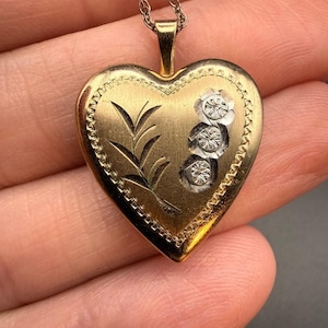 May include: A gold heart-shaped pendant featuring a textured border, an engraved leafy branch design on the left side, and three small, circular silver-toned floral accents arranged vertically on the right side, suspended from a gold chain.
