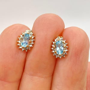 May include: A pair of stud earrings featuring oval-cut light blue gemstones set in gold metal. Each central stone is surrounded by a halo of small, clear, round-cut stones, creating a cluster design that sparkles against the gold setting.