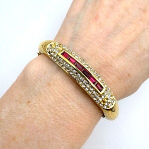 May include: Gold-toned hinged bracelet with a rectangular center design. The center features a row of rectangular, ruby-colored stones, surrounded by clear, sparkling crystals. The bracelet is worn on a wrist.