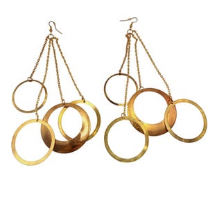 May include: A pair of gold-toned dangle earrings featuring three interconnected circular hoops of varying sizes suspended from delicate metal chains. The earrings are attached to standard fishhook wires, creating a layered, geometric design.