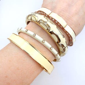 Op de afbeelding: Een set van vier gestapelde armbanden met een gladde goudkleurige bangle, een dunne crèmekleurige bangle met ingebedde heldere kristallen, een bredere goudkleurige bangle met crème emaille en kristallen accenten, en een crèmekleurige schakelarmband.