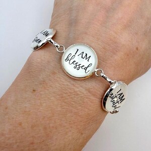 May include: Silver-toned bracelet with three circular charms. The central charm reads "I AM blessed" in black script. The other charms have similar text. The bracelet is worn on a wrist.