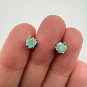 May include: A pair of stud earrings featuring heart-shaped, light green, translucent stones set in silver-toned metal prongs. Each earring has a faceted surface that catches the light, showcasing a pale mint hue and a classic heart silhouette.