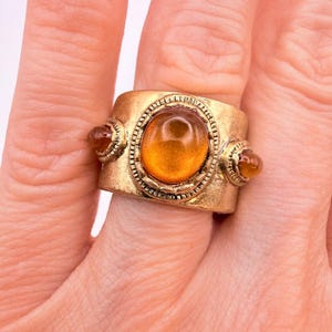 May include: A wide gold-toned metal ring featuring a large, central oval amber-coloured cabochon stone surrounded by a textured border, flanked by two smaller, matching round amber-coloured stones on either side of the band's surface.