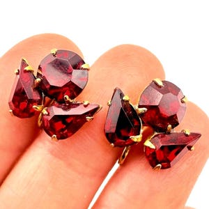 May include: A pair of earrings featuring deep red, faceted, round and pear-shaped stones set in gold-toned metal. Each earring consists of one circular stone and one teardrop-shaped stone clustered together, reflecting light through their polished surfaces.