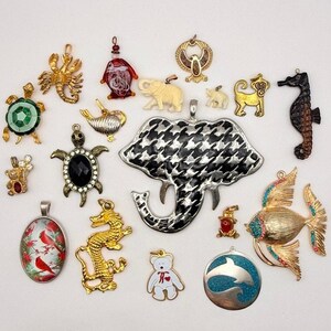 Puede incluir: Una colección de colgantes variados con temática animal en metal y esmalte, incluyendo un elefante grande con patrón de pata de gallo en blanco y negro, un dragón dorado, un caballito de mar, tortugas, un pez con pedrería azul y rosa, y un óvalo de cardenal.