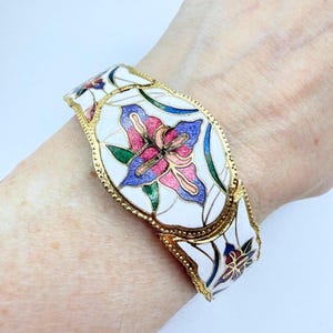 Könnte beinhalten: Ein goldfarbenes Gelenkarmband mit weißer Emaille-Basis. Das Armband hat ein Blumendesign mit rosa, blauen und grünen Akzenten. Das Design des Armbands ist symmetrisch, mit einem sich wiederholenden Muster aus Blumen und Blättern.