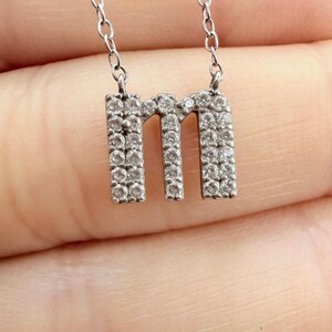 May include: A silver necklace with a pendant in the shape of the letter "m", set with small, clear gemstones. The pendant hangs from a fine chain. The necklace is shown against a neutral background.