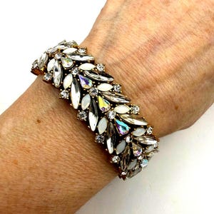 Op de afbeelding: Een goudkleurige armband versierd met een cluster van witte, heldere en iriserende kristallen. De kristallen zijn gerangschikt in een bloem- of bladachtig patroon, waardoor een sprankelend en elegant accessoire ontstaat. De armband wordt om een pols getoond.