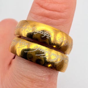 May include: Two polished gold-toned metal rings featuring an engraved script pattern that wraps around the entire outer surface of each band. The rings have a smooth, reflective finish and are stacked vertically, displaying the intricate lettering.