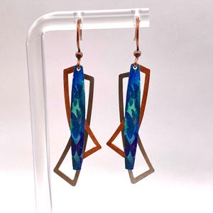 May include: Dangle earrings with a geometric design. Each earring has a blue and green patterned central element, a copper-coloured frame, and a silver-coloured geometric shape. The earrings hang from copper-coloured hooks.