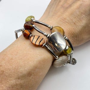 Puede incluir: Pulsera de múltiples hilos con barras de metal plateado y una variedad de cuentas. Las cuentas incluyen formas ovaladas, redondas y de disco en tonos de marrón, naranja, amarillo y gris. La pulsera se usa en la muñeca.