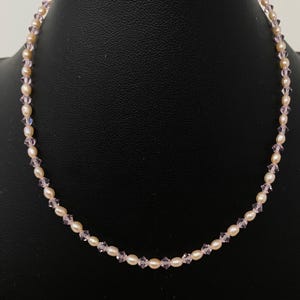 May include: A delicate necklace featuring small, creamy-white pearls interspersed with sparkling, light-pink crystals. The necklace is displayed against a dark background, highlighting its elegant design. This piece is a classic jewelry item.