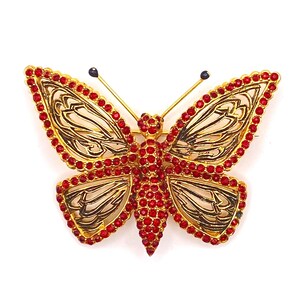 Peut inclure: Une broche papillon dorée avec un corps central et des bords d'ailes incrustés de petits strass rouges. Les ailes présentent des détails en filigrane ajouré en métal doré, et la tête comporte deux fines antennes aux extrémités sombres.