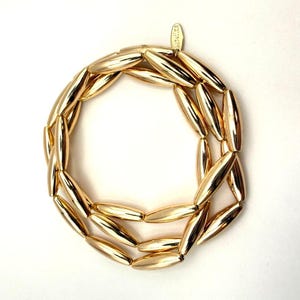 Op de afbeelding: Een goudkleurige ketting bestaande uit langwerpige, ovaalvormige kralen. De kralen zijn aan elkaar geregen tot een doorlopende lus. Een klein label is bevestigd bij de sluiting. De ketting wordt getoond tegen een witte achtergrond.