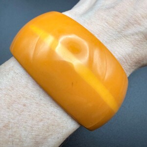 May include: A wide, smooth, solid orange bangle bracelet with a glossy finish. The bracelet features a subtle, carved diagonal pattern across its surface, creating a uniform, rounded shape that encircles the wrist with a bright, vibrant color.