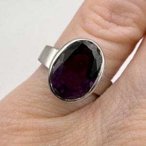 May include: A silver ring featuring a large, oval-cut, deep purple gemstone. The gemstone is set in a bezel setting, and the band is a simple, wide design. The ring is shown on a person's finger.