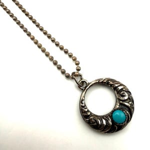 Op de afbeelding: Een zilverkleurige hangerketting met een cirkelvormig, sierlijk ontwerp. De hanger heeft een heldere turkooizen steen en hangt aan een kogelketting. De ketting wordt gepresenteerd tegen een witte achtergrond.