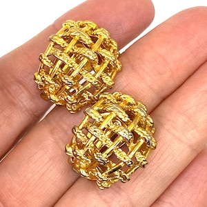 Può includere: Un paio di orecchini dorati, in stile intrecciato. Gli orecchini hanno un design arrotondato a forma di gabbia, con una superficie strutturata. Gli orecchini sono mostrati su uno sfondo color carne.