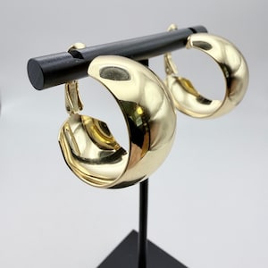 May include: A pair of gold-colored, clip-on hoop earrings. The earrings are displayed on a black stand against a white background. The hoops have a rounded, polished design, reflecting light.