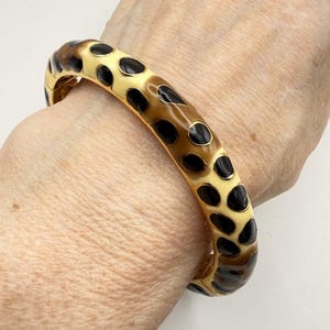 May include: A hinged bangle bracelet featuring a glossy enamel finish with a repeating pattern of black spots against a cream and light brown gradient background. The bracelet has a smooth, rounded profile and a metallic gold-tone interior frame.