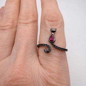Peut inclure: Bague noire en forme de chat, ornée d'une pierre rouge en forme de goutte. La queue de la bague s'enroule autour du doigt, et le corps du chat est orné de petites pierres noires. Sur fond neutre.