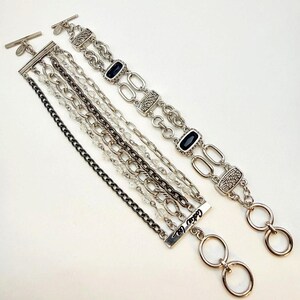 May include: Two silver-toned bracelets with toggle clasps. One features multiple layered chains with clear beads and a bar engraved with Cookie Lee. The other consists of oval links, textured rectangular segments, and two dark rectangular stones.