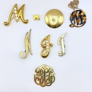 May include: A collection of vintage monogram jewelry pieces, including brooches, a pendant necklace, and a small pin. The pieces are in gold and silver tones, with some featuring rhinestones. The jewelry is arranged on a white surface.