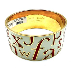 May include: A gold-tone bangle bracelet with a light gray enamel band featuring red letters. The inside of the bracelet is gold. The text "kate spade NEW YORK" is visible.