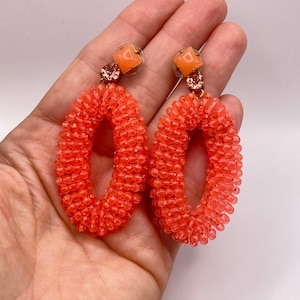 Op de afbeelding: Een paar koraalkleurige, met kralen bezette ovale oorbellen met een vierkante oranje knop aan de bovenkant, geaccentueerd met een kleine ronde gefacetteerde kristal. De ovale hangers zijn opgebouwd uit talloze kleine, doorschijnende koraalkralen.