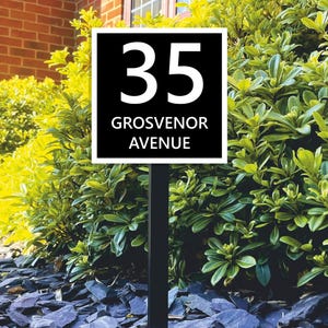 Könnte beinhalten: Ein schwarz-weißes Adressschild mit der Nummer "35" und "GROSVENOR AVENUE". Das Schild ist an einem schwarzen Pfosten befestigt und in einem Gartenbeet mit dunkelgrauen Steinen und grünem Laub angebracht.